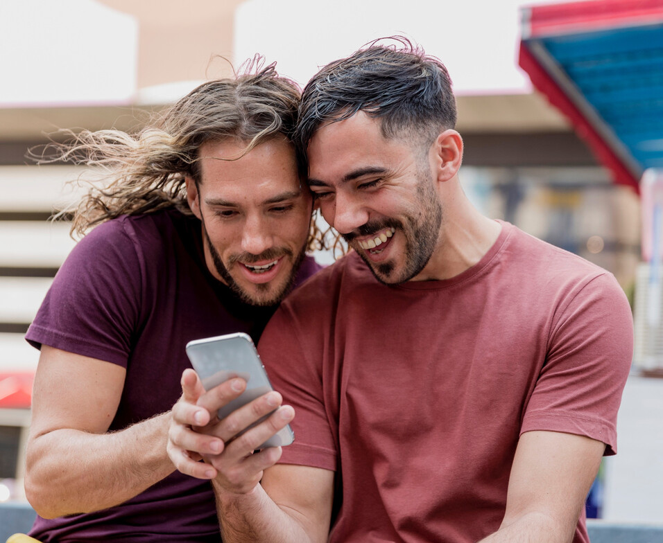 An LGBTQIA couple is sitting on a stoop and looking at a phone to learn more about online couples therapy